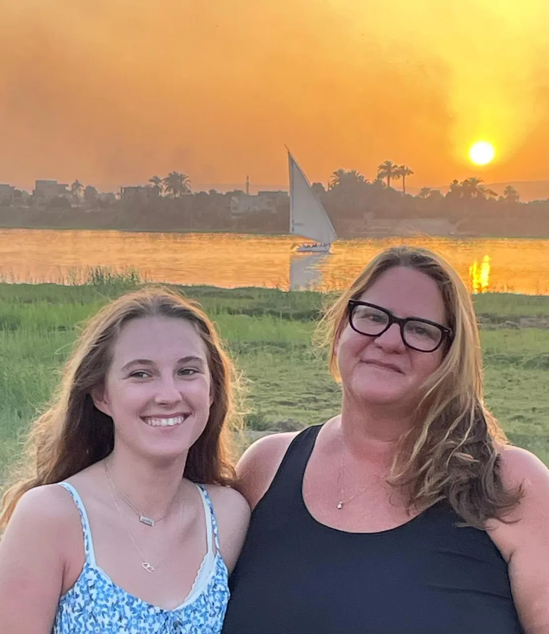 Two women smiling by the Nile at sunset, with a felucca behind them